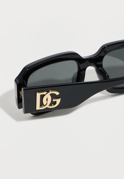 Μαύρα ορθογώνια γυαλιά ηλίου με σκούρους φακούς και χρυσά αρχικά που ενώνονται στο χοντρό πλευρικό μπράτσο, πάνω σε λευκό φόντο.