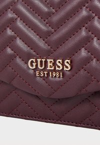 Bordeaux, kviltet, kunstlæder håndtaske med guld "GUESS EST 1981" logo, der har et chevronmønster og kontrasterende syninger.