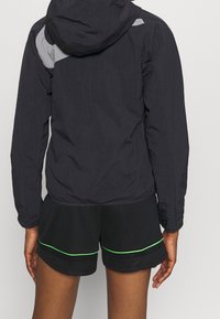 Black lightweight jacket with a grey panel on the back, featuring a hood, elastic cuffs, and paired with black shorts with green accents.