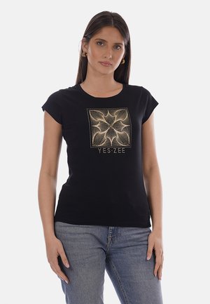 Donna che indossa una T-shirt nera aderente con un motivo geometrico dorato e la scritta "YES ZEE", abbinata a jeans di denim blu.
