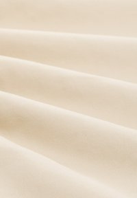 Tessuto di cotone in una leggera tonalità beige, con texture morbide e ripiegate che creano ombre e profondità sottili. Nessun motivo o hardware visibile.