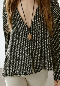 Kobieta nosząca czarny i biały sweter o fakturze w serek z długimi rękawami oraz brązowy naszyjnik z wisiorkiem na jasnych spodniach.