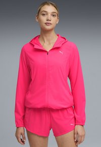 Γυναίκα που φοράει φωτεινό ροζ φούτερ Puma με φερμουάρ και ταιριαστό σορτς, στέκεται μπροστά σε ουδέτερο γκρίζο φόντο με διαβαθμίσεις.