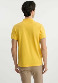 Żółta koszulka polo z krótkim rękawem, z kontrastującymi szarymi akcentami na mankietach, wykonana z gładkiego, lekkiego materiału, pokazana z tyłu.