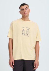 T-shirt jaune en coton à manches courtes, avec une impression graphique de deux personnages assis. Coupe décontractée avec un col rond.