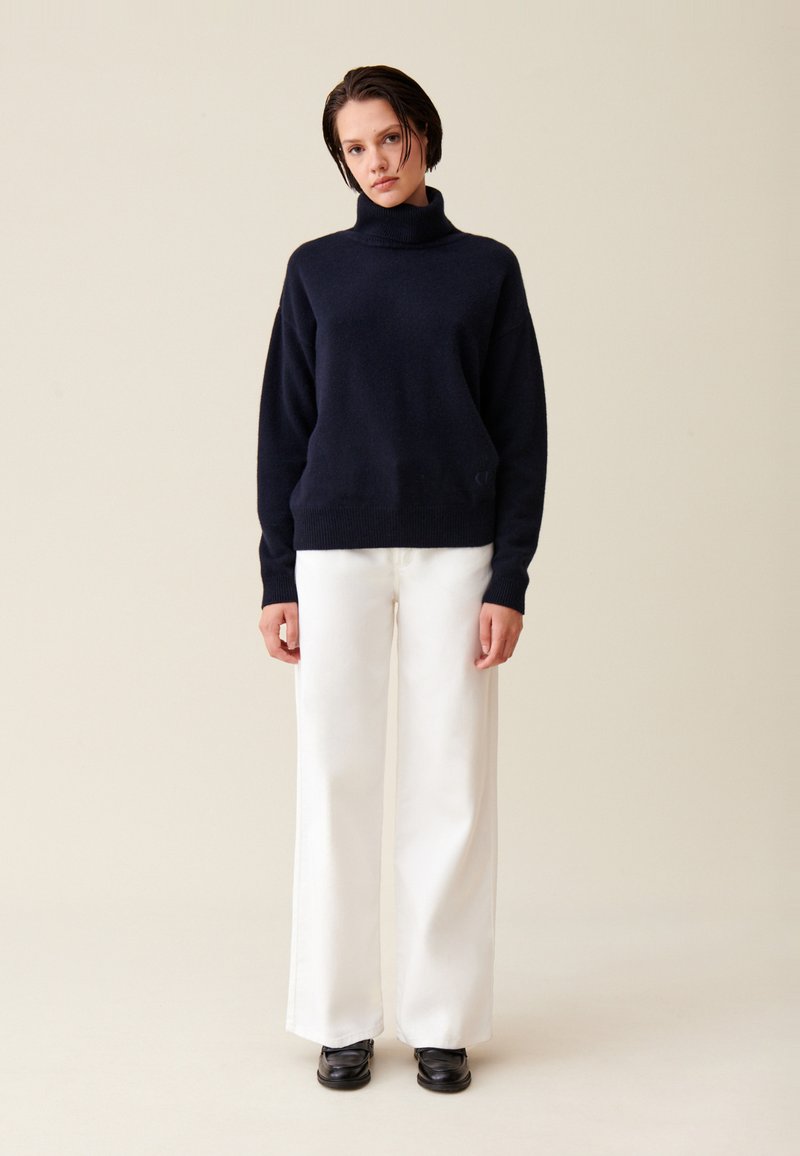 Pulover din tricot cu guler înalt de culoare marine, asortat cu pantaloni albi cu picioare largi și pantofi negri. Textură netedă, croială lejeră, branding minim.