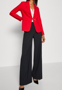 Rød skræddersyet blazer med skarpe klapper, kontrasterende sorte bredde ben-bukser og højhælede sorte sko, med et elegant design og en glat tekstur.