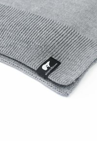 Beanie in maglia grigia con design a coste. Presenta un'etichetta nera con logo e testo bianchi. Texture morbida con dettaglio di orlo piegato.