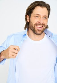 Lyseblå skjorte med knapper, båret åben over en almindelig hvid t-shirt, med korte ærmer og en glat tekstur. Mand der smiler.