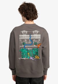 Sudadera gris con un gráfico colorido en la parte posterior que muestra un edificio con plantas y animales, contorneado en blanco y acentuado con azul, naranja y púrpura.