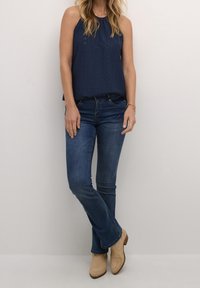 Kvinna i en ärmlös marinblå topp, åtsittande blå jeans, beige ankelstövlar och ett guldarmband som står mot en enkel vägg.