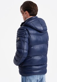 Granatowa kurtka puchowa z kapturem, pikowana, z elastycznymi mankietami i teksturowanym wykończeniem. Posiada naszywkę z logo na lewym ramieniu.