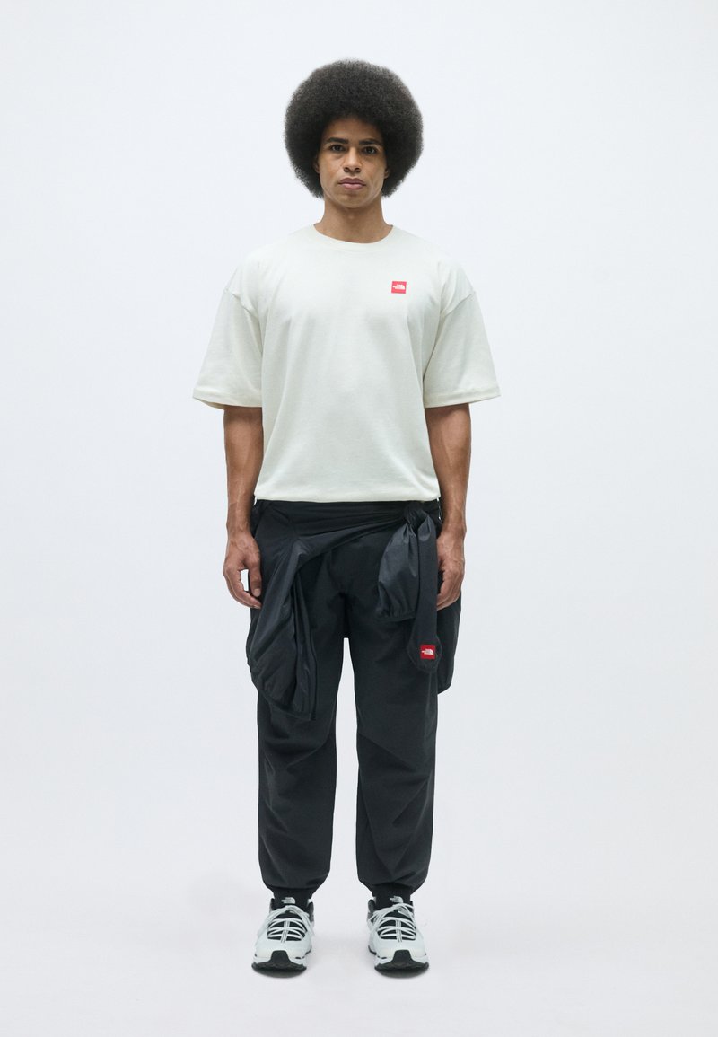 Ljust grå oversized t-shirt med en liten röd logotyp, parat med svarta byxor med dragsko och fickor. Svart och vita sneakers.