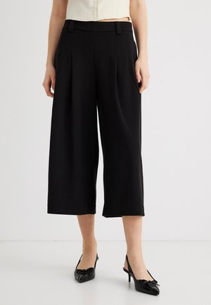 Femme portant un pantalon large noir raccourci, des escarpins noirs à bride arrière, et un top blanc raccourci boutonné, debout sur un sol blanc.