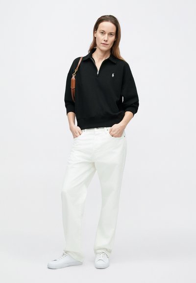 Svart genser med glidelås og logo, hvite bukser med høy midje og hvite joggesko, komplettert med en brun veskerem.