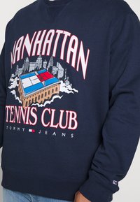 Felpa marina con grafica grande "Manhattan Tennis Club" che presenta un design di un campo da tennis, accenti rossi e blu, e polsini a costine.