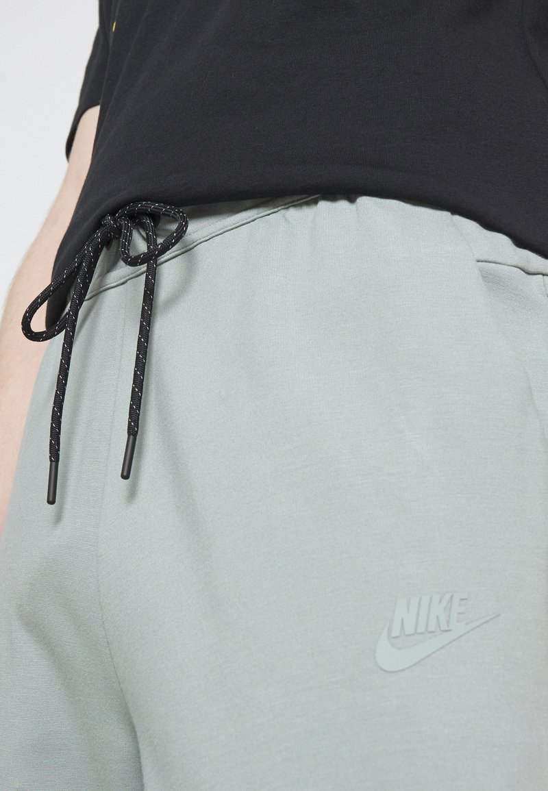 Gros plan sur une personne portant un pantalon de sport Nike gris clair avec cordon noir et un t-shirt noir.