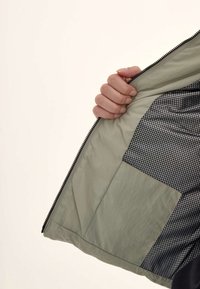 Una mano sosteniendo abierta una chaqueta para revelar un forro verde claro con un interior de patrones en blanco y negro, mostrando bolsillos y texturas.