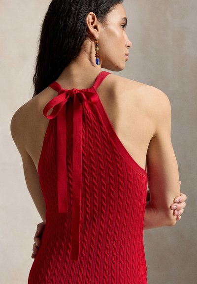 Femeie cu păr lung și închis la culoare poartă un top halter tricotat roșu legat cu o fundă la gât, care își lasă umerii și spatele goale, purtând o cercească colorată și pendulantă.