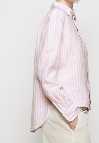 Ljust rosa och vit randig knytskorta med krage, långa ärmar och rundad nederkant. Tillverkad av ett strukturerat tyg.