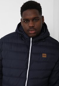 Marineblå pufferjakke med vandret syninger. Har en hvid lynlås, justerbar hætte og et brunt læderlogo-patch på brystet.