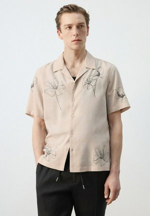 Joven con una camisa beige de manga corta con dibujos florales negros y pantalones negros, de pie frente a un fondo liso.
