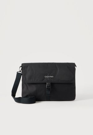 Sac messager noir Calvin Klein avec bandoulière réglable et rabat avant sécurisé par une boucle, présenté sur un fond uni.