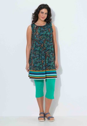 Mujer con cabello rizado que lleva una túnica sin mangas de flores con dobladillo a rayas, leggings verdes recortados y sandalias azules, de pie contra una pared blanca.