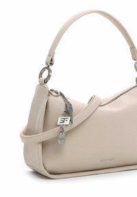 Beige leren handtas met een korte handgreep, een zilveren ritssluiting en decoratieve zilveren bedeltjes die dicht bij de handgreep hangen.