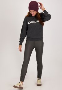 Grijze sweatshirt met de tekst "Couture", slim olijfgroene broek en beige sneakers met maroon accenten, afgewerkt met een bordeauxrode gebreide beanie.