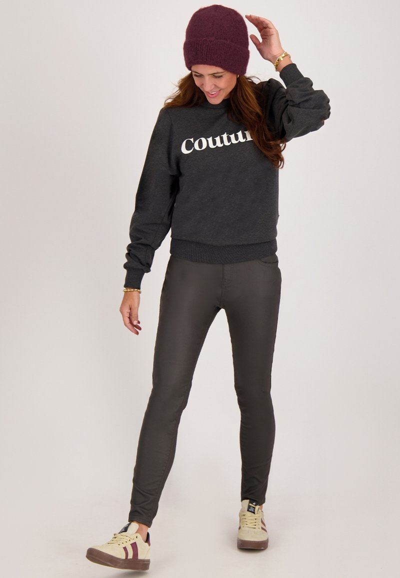 Grijze sweatshirt met de tekst "Couture", slim olijfgroene broek en beige sneakers met maroon accenten, afgewerkt met een bordeauxrode gebreide beanie.