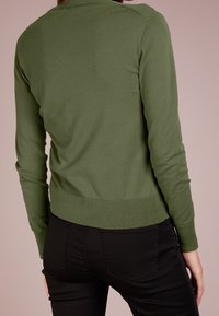 Olivgrön långärmad tröja med en figurnära design och ribbad nederkant, tillverkad av mjukt stickat material. Buren med svarta jeans, bakvy.