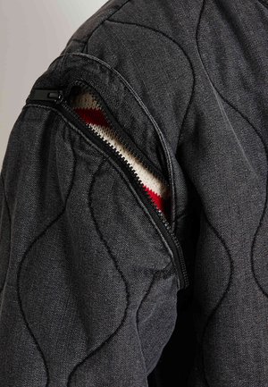 Veste matelassée noire avec une manche gauche zippée révélant un intérieur en tricot avec des rayures rouges et blanches. Tissu structuré et texturé.