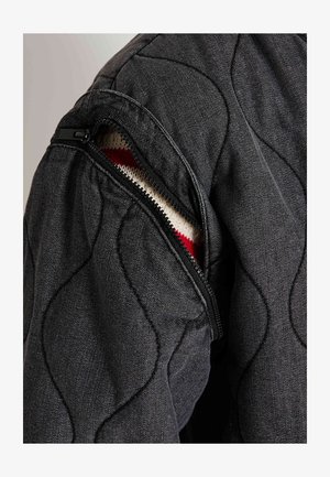 Chaqueta negra con acolchado que presenta una manga izquierda con cremallera, revelando un interior de punto con rayas rojas y blancas. Tejido estructurado y texturizado.