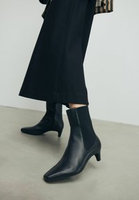 Schwarze Stiefeletten haben eine spitze Zehenform, ein glattes Obermaterial aus Leder, elastische Seiteneinsätze und einen niedrigen Absatz. Das Design steht im Kontrast zu einem schwarzen Rock.