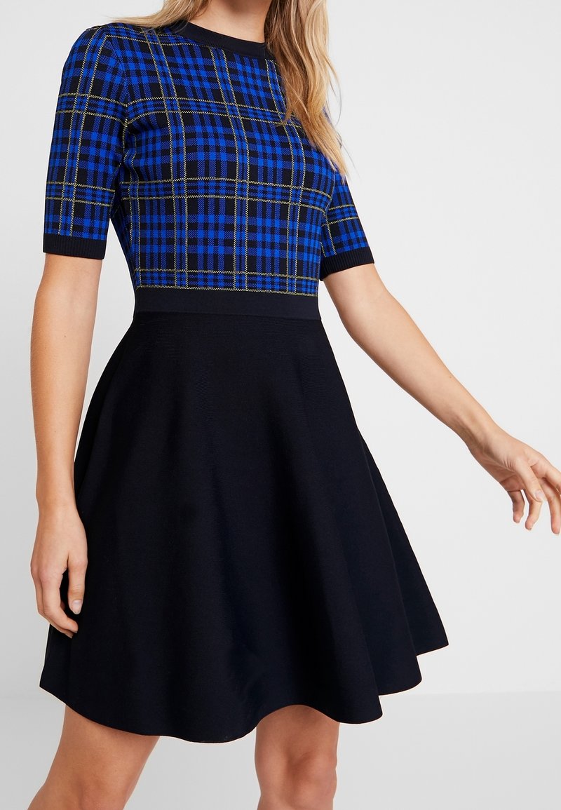 Femme portant une robe à manches courtes avec un haut à carreaux bleu et noir et une jupe évasée unie noire, sur un fond uni.