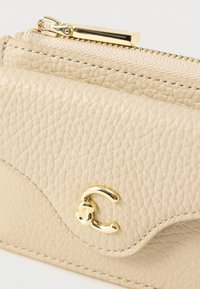 Μπεζ δερμάτινο πορτοφόλι με υφή, χρυστό φερμουάρ και λεπτομέρεια λογότυπου. Διαθέτει κυρτή άκρη και ορατή ραφή.