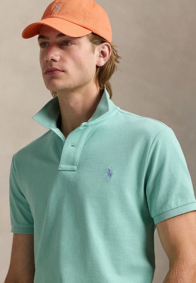 Nuori mies, jolla on oranssi lippis ja mintunvihreä pikeepaita nostetulla kauluksella, katsoo vasemmalle yksiväristä beigeä taustaa vasten.