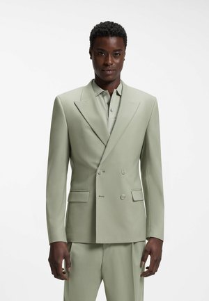 Man draagt lichtgroene dubbelgeknoopte blazer en bijpassende broek over een overhemd met kraag, staand tegen een witte achtergrond.