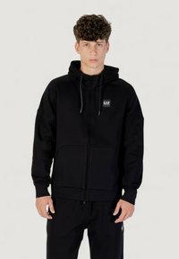 Schwarze Zip-Up-Kapuzenshirt aus weichem Material, mit Kapuze und Kordeln, Fronttaschen und einem kleinen Logo-Patch auf der Brust.