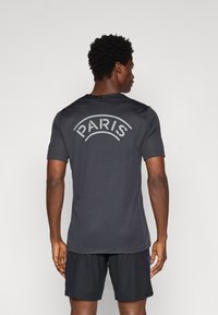 Czarna sportowa koszulka z krótkim rękawem, z napisem "PARIS" w zakrzywionym białym tekście na górnej części pleców, noszona z dopasowanymi czarnymi szortami.