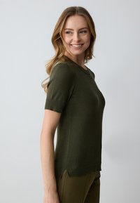 Lächelnde Frau mit langen, welligen blonden Haaren, die einen dunkelgrünen, kurzärmeligen Pullover und passende Hosen trägt und über ihre Schulter schaut.