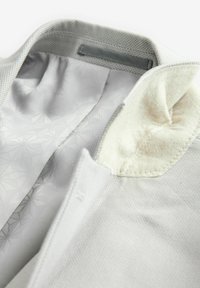 Interior de una chaqueta gris claro con una tela texturizada y un patrón geométrico, forro de cuello en color crema suave y detalles de costura visibles.