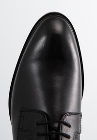 Chaussure habillée en cuir noir avec une surface lisse et brillante, un bout arrondi et un système de laçage, ornée de détails texturés sur les côtés.