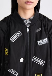 Melna bomber jaka ar dažādiem kuģiem, tostarp "DKNY EST. 1989" un "DON'T WALK." Iezīmes ietver rāvējslēdzēja priekšpusi un ribotiem apkakli.