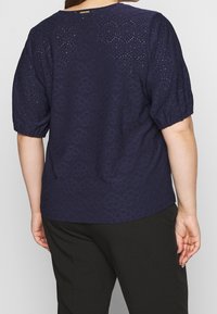 Námořnická modrá blůza s texturovaným květinovým vzorem, krátkými nabíranými rukávy a kulatým výstřihem, doplněná tmavými černými kalhotami.