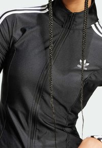 Svart sportjacka med hög krage, dragkedja fram och vita tre-randiga detaljer längs axlarna, med en broderad logotyp på bröstet.