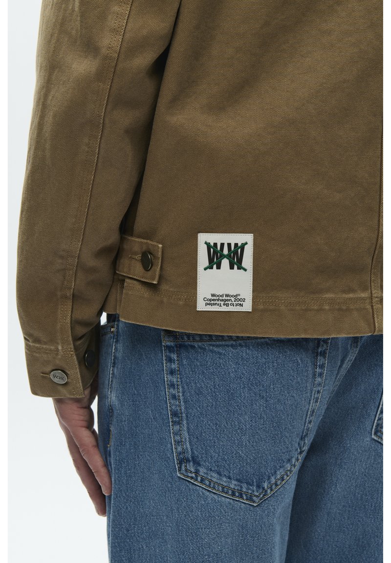 Veste en toile marron avec une étiquette, dotée d'un poignet boutonné et d'une coupe décontractée. Associée à un jean en denim bleu, montrant le détail de la poche arrière.