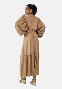 Robe midi marron avec des manches bouffantes, une taille cintrée et une jupe à volants. Le mannequin porte des sandales compensées, mettant en valeur un tissu fluide et un design simple.