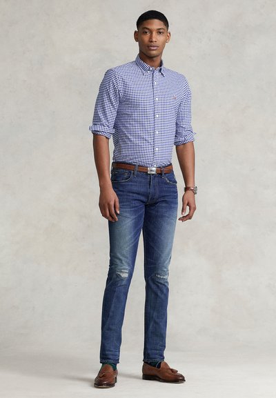 Ung mand stående iført blå og hvid ternet skjorte med opsmøgede ærmer, blå jeans med slidte knæ, brun bælte og sko.